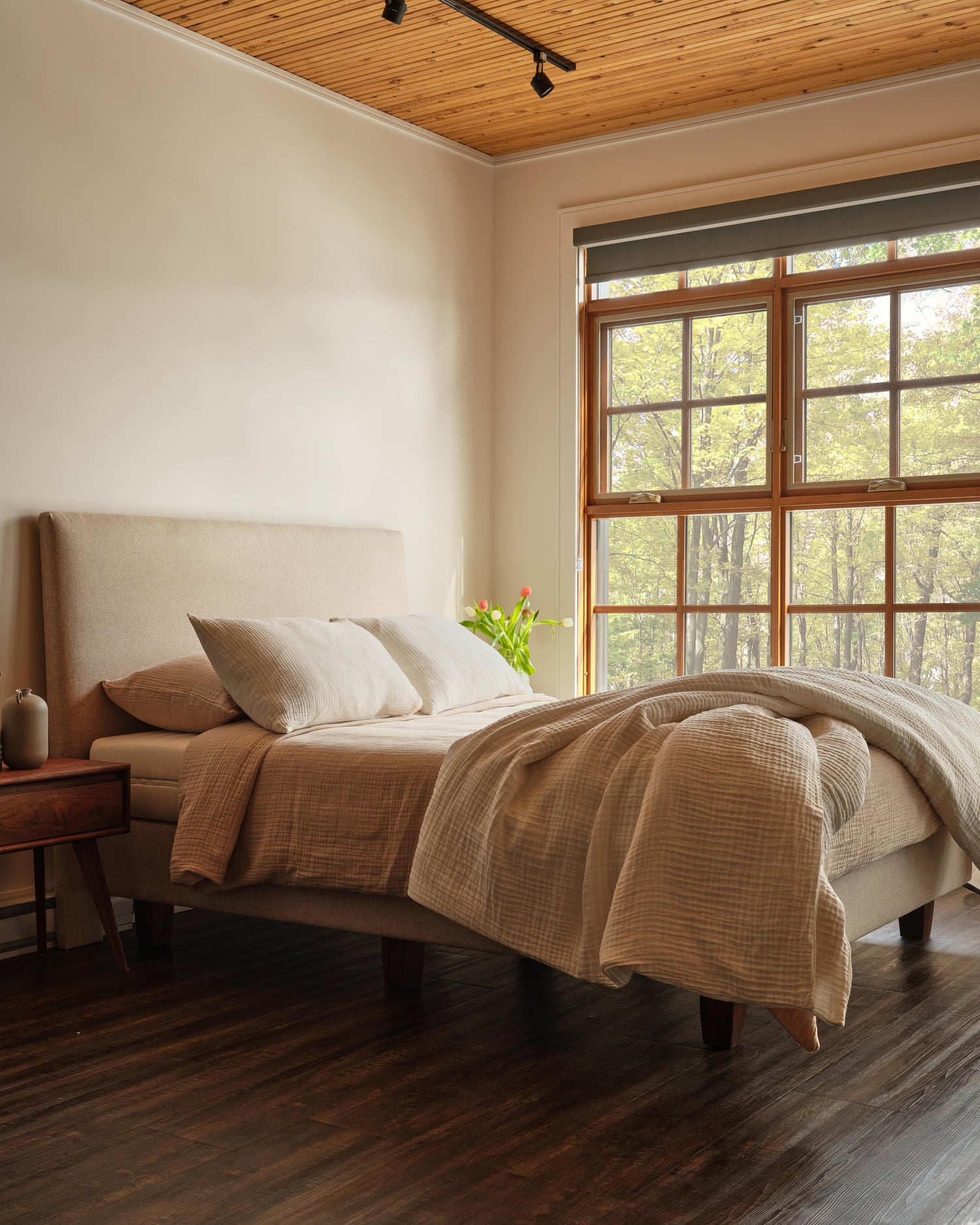 Bedroom with a large window view of trees, beige bedding, and wooden ceiling.