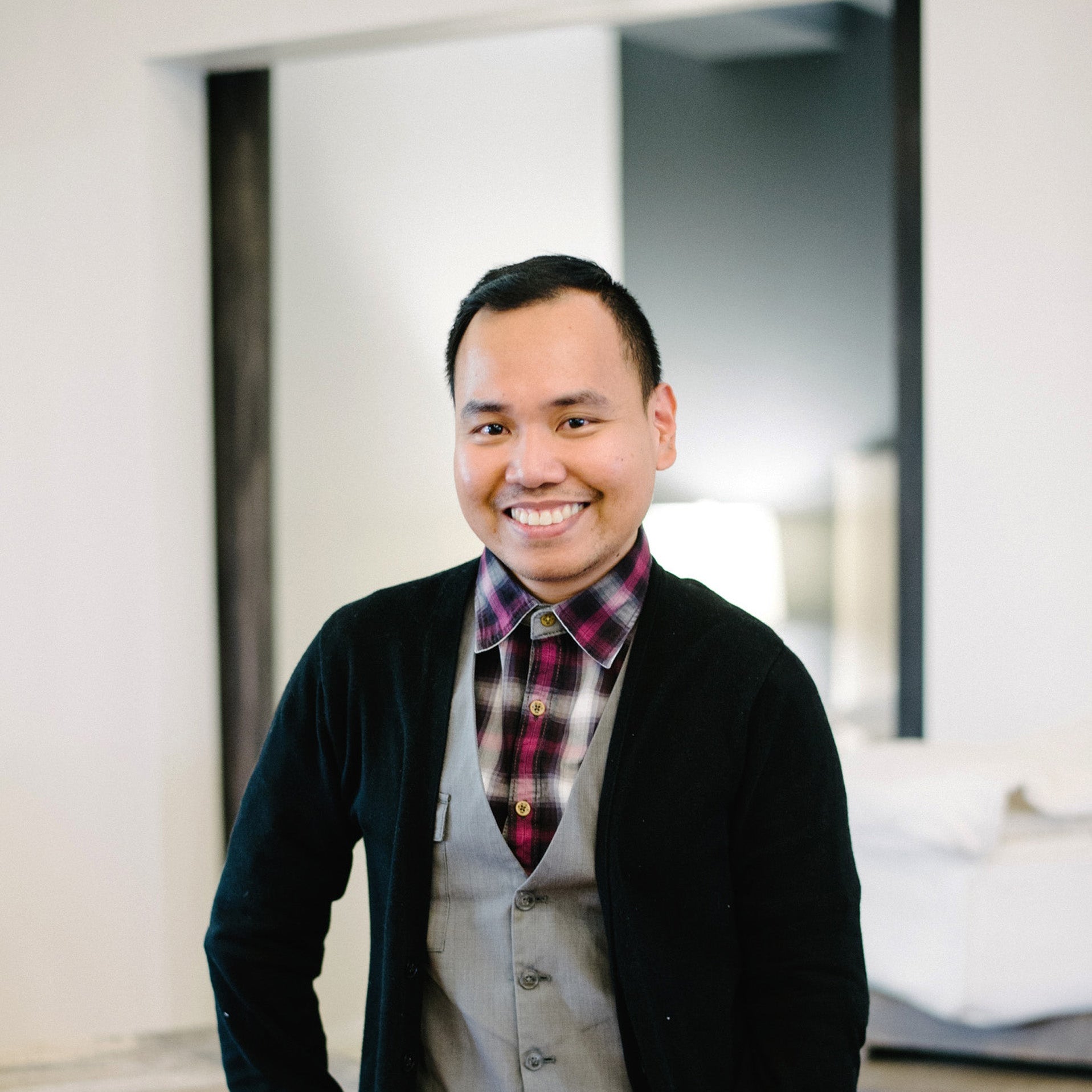 Man wearing a black cardigan, plaid shirt, and vest indoors.