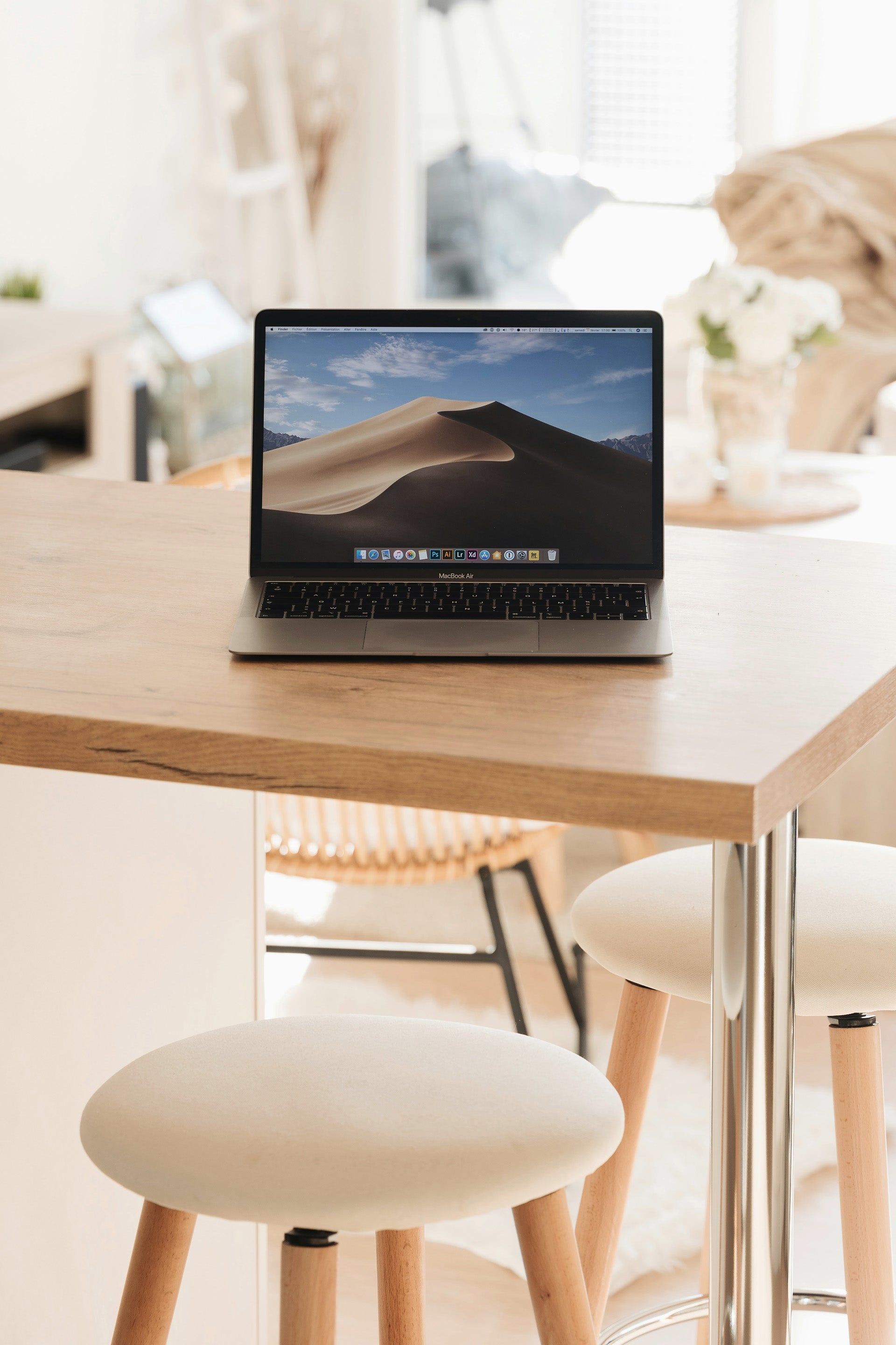 Laptop on a wooden table with a desert landscape on the screen, surrounded by chairs and a blurred indoor setting.